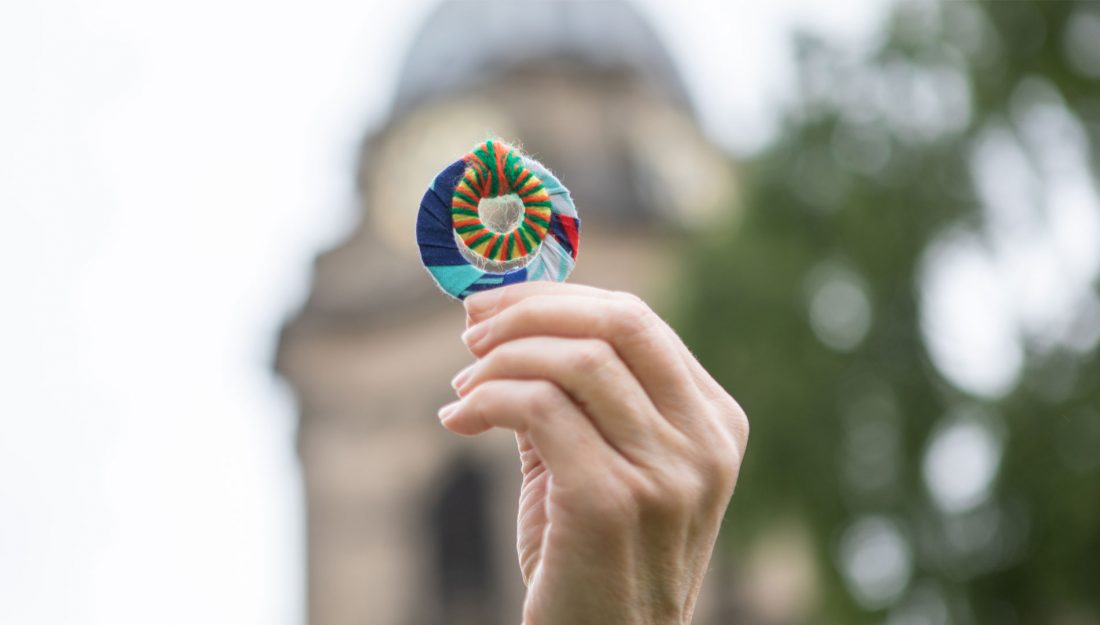 a hand holding up a handmade textile object made up of two rings wrapped in colourful yarn with a cathedral in the background