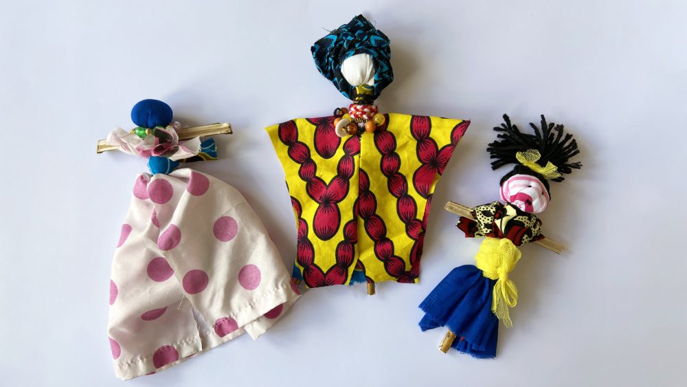 Three stick dolls made from colourful fabrics and adorned with beads rest on a white background