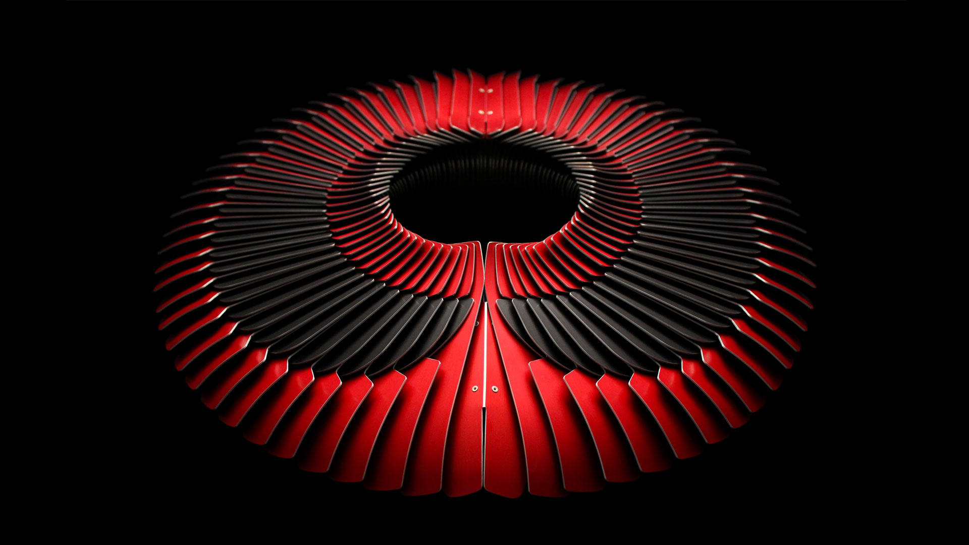 A large circular red and black metal neckpiece constructed like a collar and photographed on a black background. There are three layers of flat and shaped metal segments which overlap.