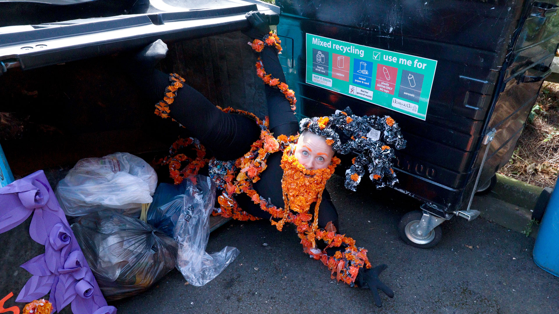 Kitt, white human coming out of an industrial size bin with arms outstretched. They are wearing a headdress, beard and garlands of flowers handmade from recycled black and orange plastic bags. Behind their head is another bin with a vinyl sign which reads mixed recycling and pictures of items that can be put in the bin.