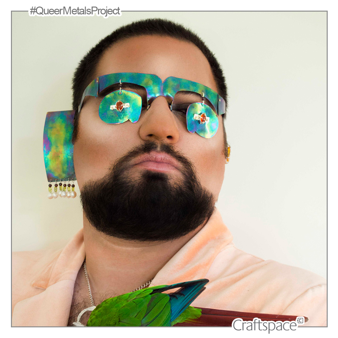 Rainbow hued non-functional glasses are worn by a person with black hair and beard, wearing contour makeup. Another rainbow hued rectangle extends from the side of the face and a green parrat perched on the lapel of a pink top.