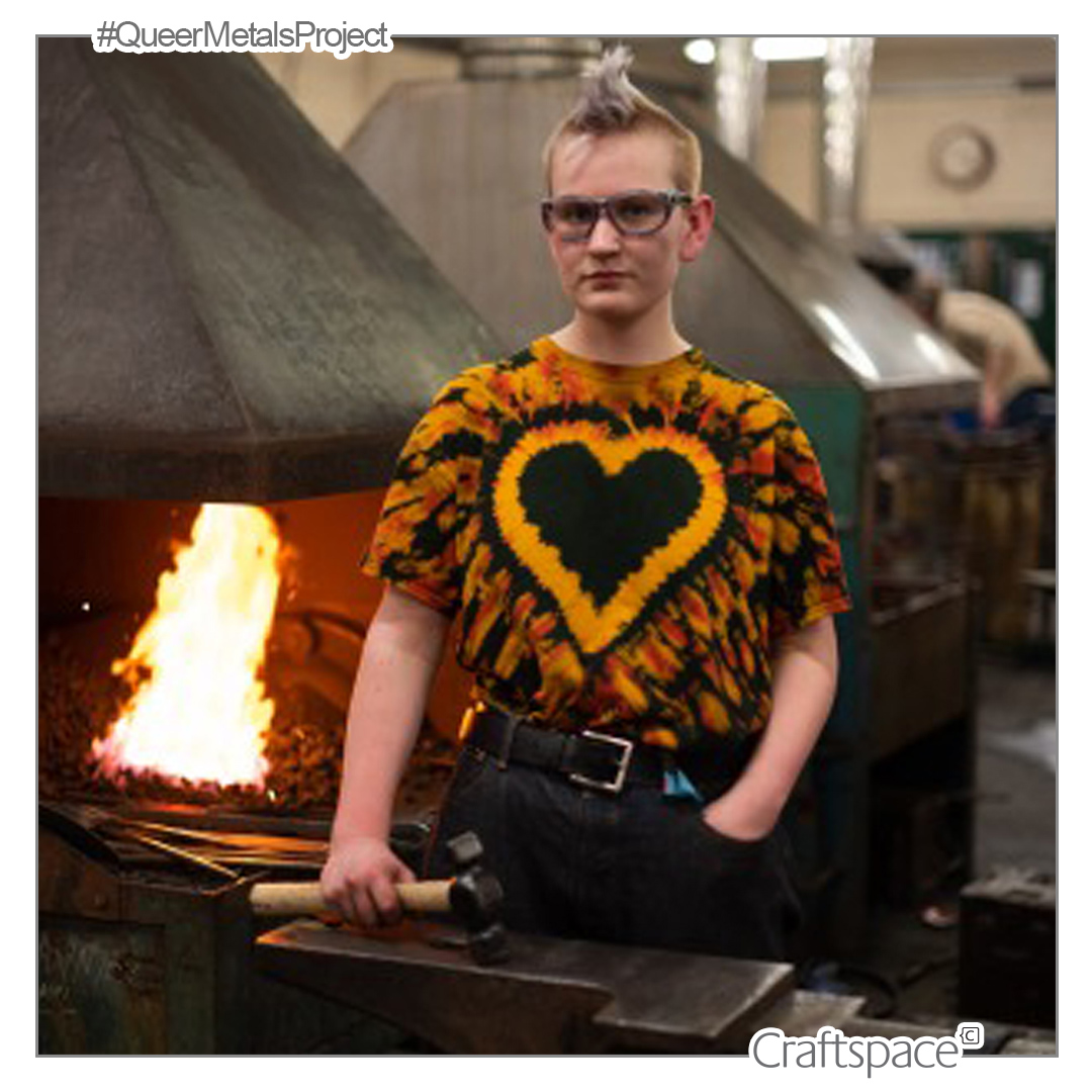 a person with blonde hair wears glasses and a tie die heart t-shirt, stands in front of a lit coal forge holding a hammer.