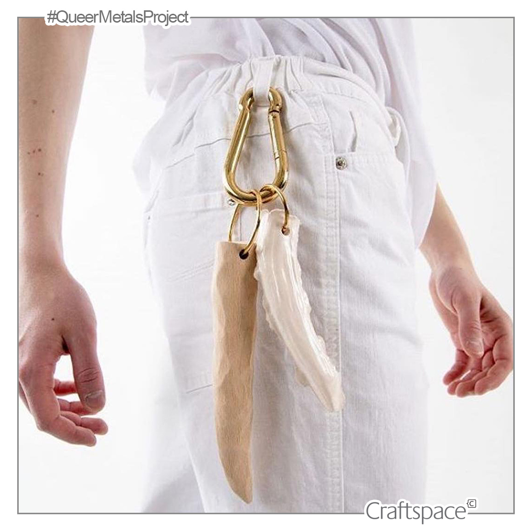 a figure seen from the waist down wearing all white with a brass carabiner on the hook of their jeans which holds two elongated objects in white and natural wood colour.