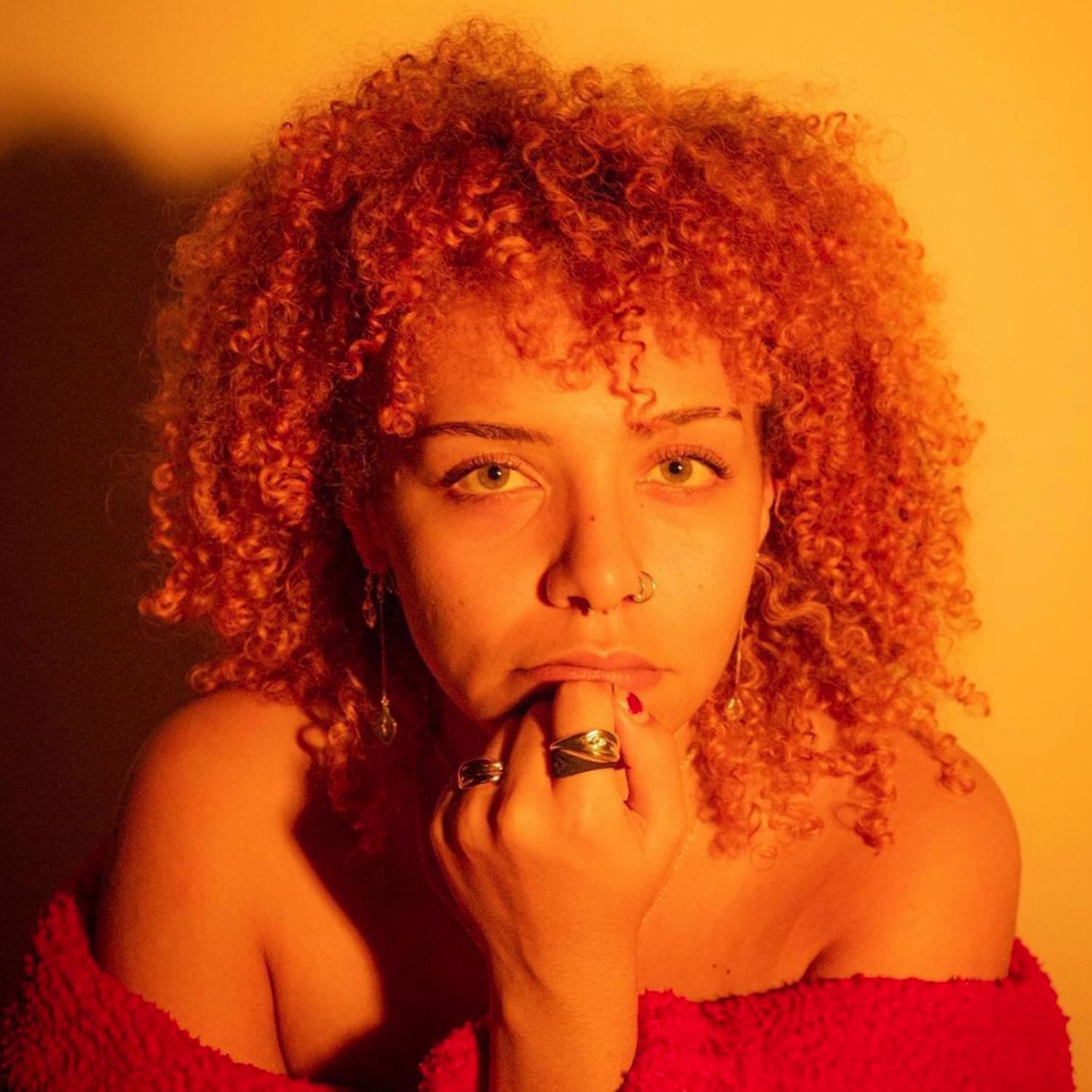 Against a yellow background, a person rests their chin on their fist while looking directly into the camera; they have curly hair, brown skin and are wearing an off the shoulder fleece