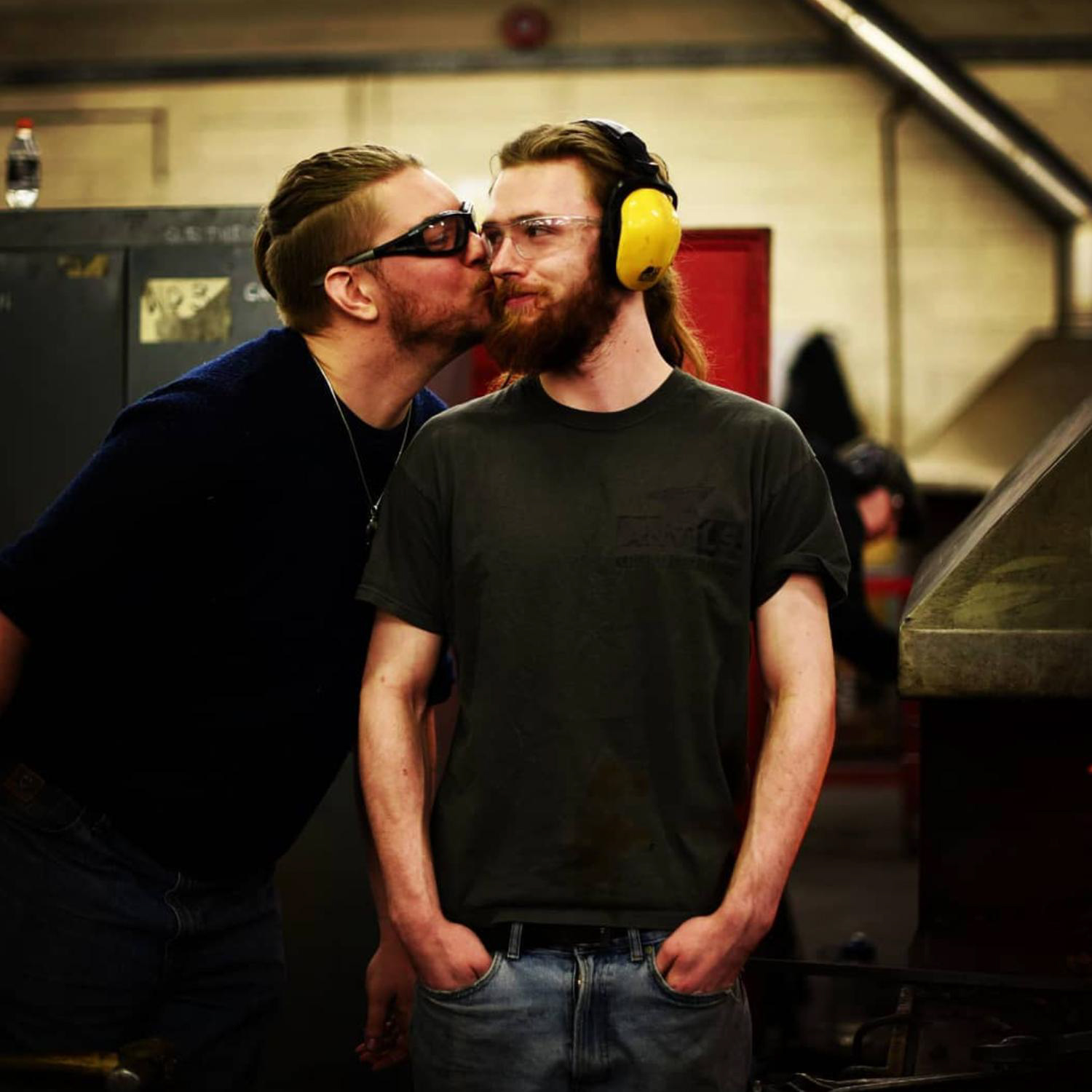 Two bearded figures wearing safety glasses and dark shirts stand in a workspace, one leans in to plant a friendly kiss on the other's cheek