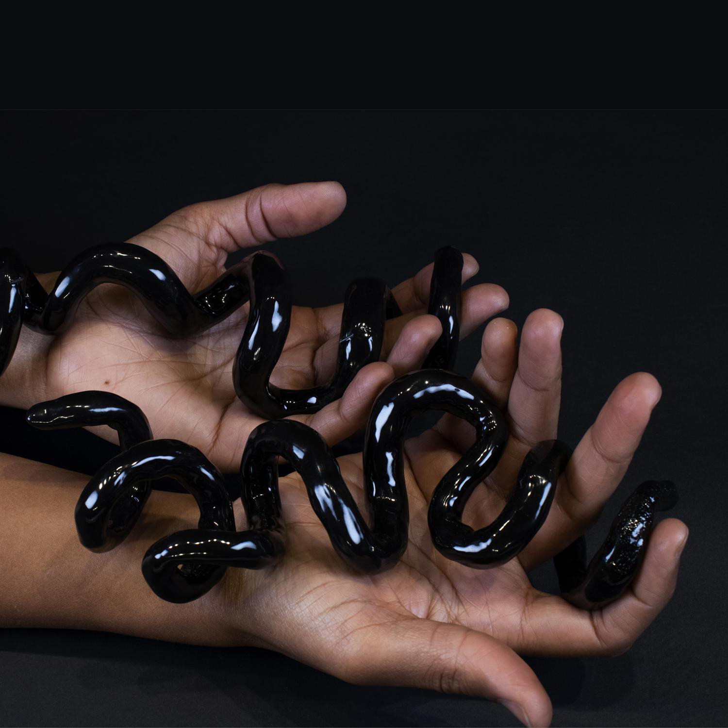 A pair of brown hands cradle shiny black squiggles that curl around fingers against a black background