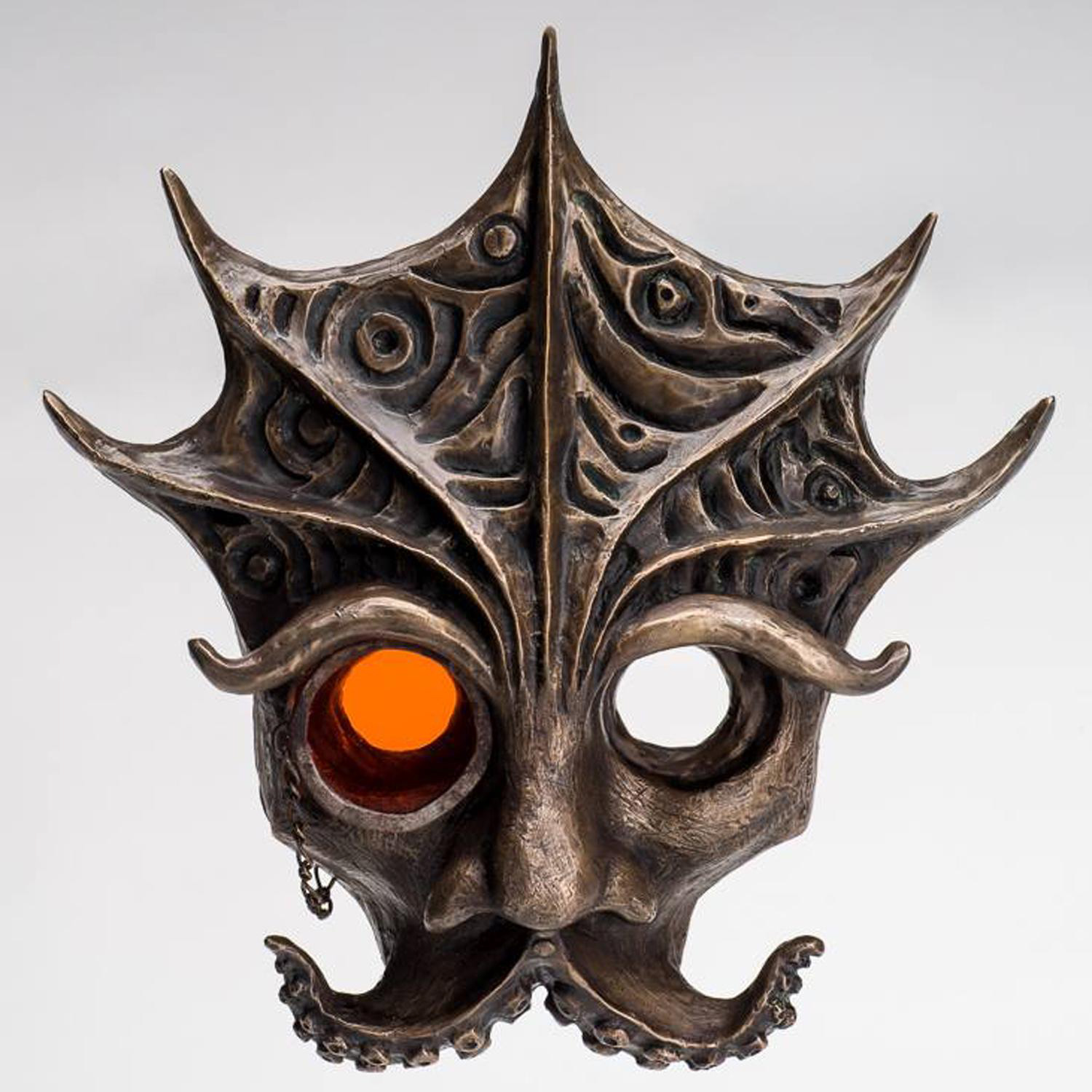 A metal sculptural face with octopus tentacles forming a mustache, broad nose, and expressive eyebrows; the forehead has deep carvings and ridges gracefully extending upwards; one eye has an orange lensed monocle with chain