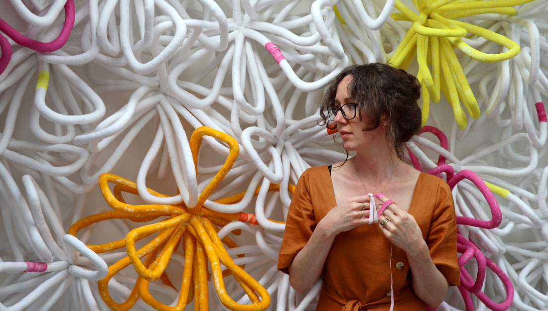A person dress in an orange dress stands against a wall of large abstract textile flowers.