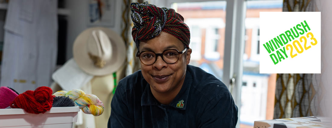 a Black woman rests her elbows on a table in a studio and looks at the camera. There are spools of thread, a box of yarn and other textiles and tools around her.