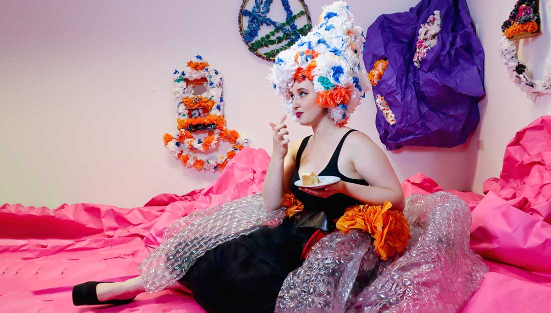 Colour photo, interior. A white Drag performer sits on the floor on a giant pink piece of textured paper. They are wearing a giant bubble wrap poofed out skirt with orange decorative plastic flowers on the top, a black bralette top and have a giant wig made out of blue, white and orange plastic flowers. They are holding a piece of cake on a saucer and bringing a finger towards their mouth.