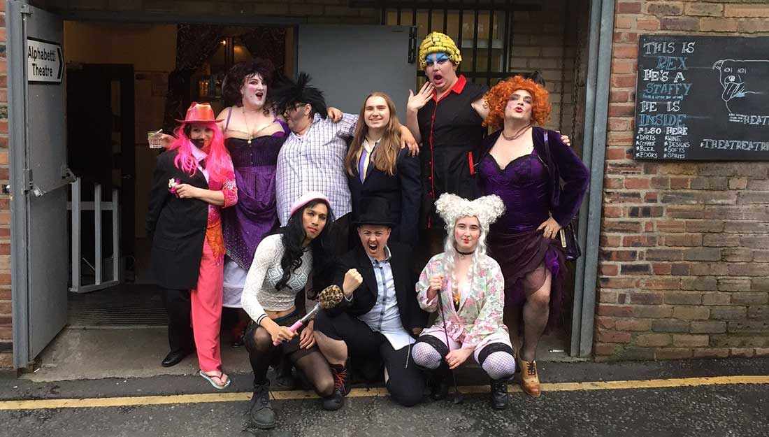 Colour photograph, exterior. A group of 9 Drag performers all posing in the doorway to a theater. There are lots of wonderful wigs and costumes and fabulous poses.