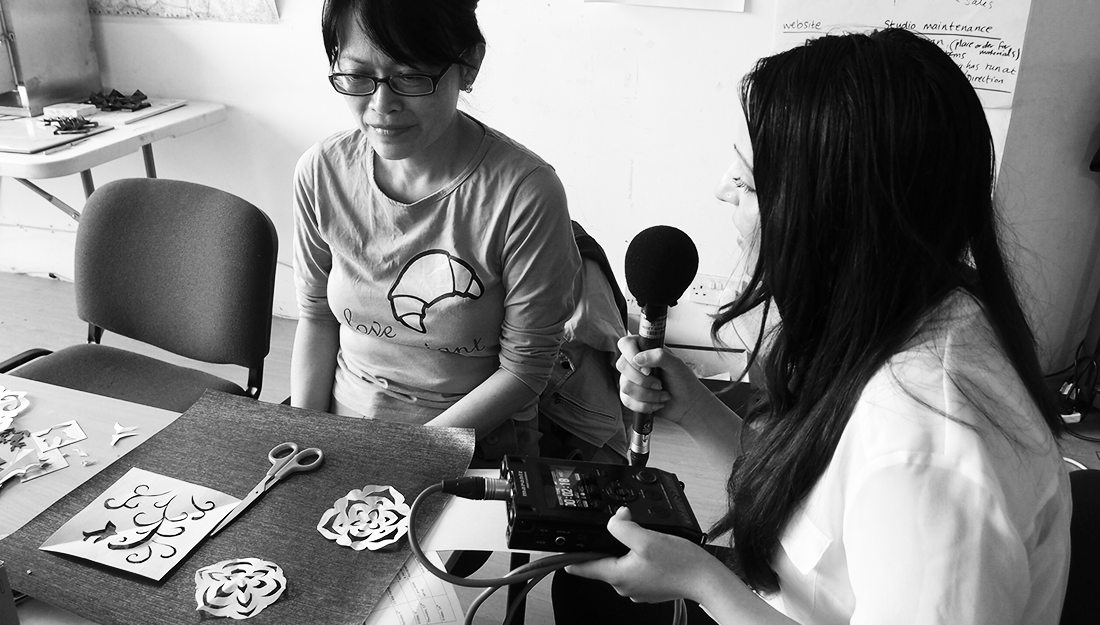 A middle aged chinese woman sits at a table in front of simple paper cuts. Beside her an interviewer holds a microphone and recording device.