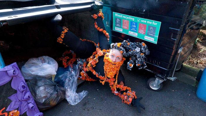A person lies on the ground as if falling out of a large overturned bin full of bin bags. They are dressed fully in black clothes decorated with strings of orange plastic flowers, a beard made from the same orange flowers and a decorative hat of black and orange plastic.