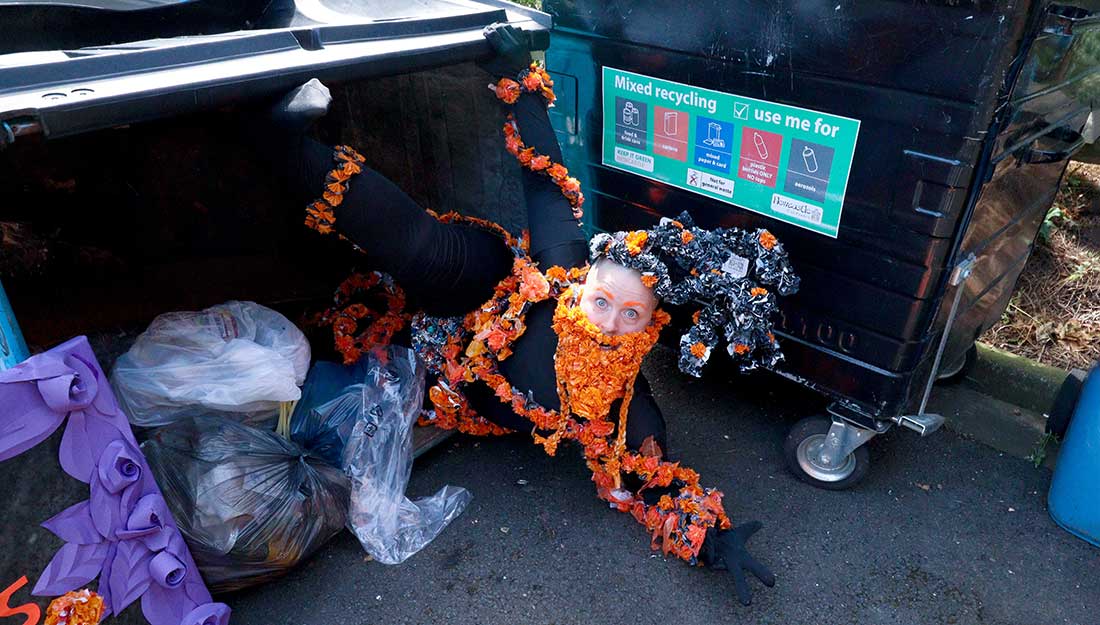 A person lies on the ground as if falling out of a large overturned bin full of bin bags. They are dressed fully in black clothes decorated with strings of orange plastic flowers, a beard made from the same orange flowers and a decorative hat of black and orange plastic.