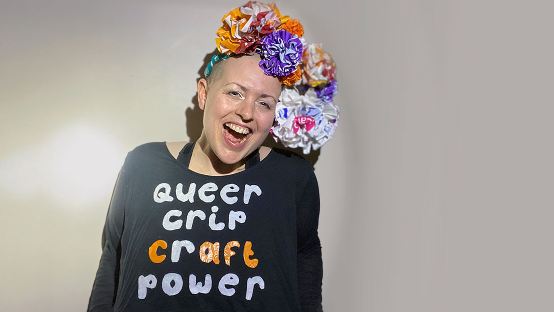 Colour photo, interior. Kitt, white shaven headed human with a big smile wears a headdress made of huge multi coloured flowers and a black long sleeved shirt on which white and orange text reads “queer crip craft power”.