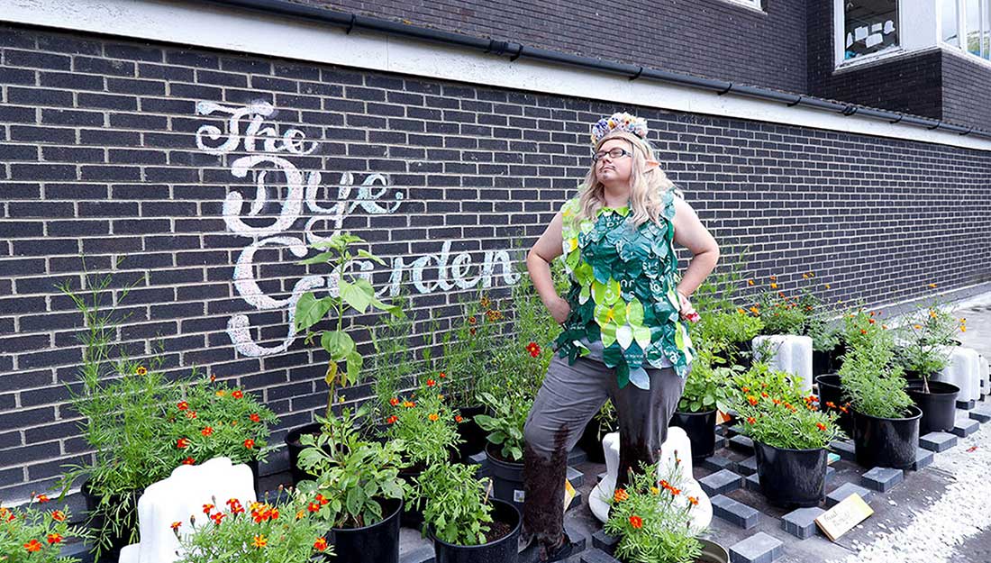 colour photo, exterior. A white Drag performer stand amidst a garden of orange and red potted flowers. They wear a leafey tabard made of green, platsic shopping bags and trousers which seems to be partly made from mud. behind them white chalk lettering on a brown brick work wall reads "The Dye Garden".