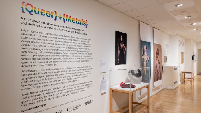 colour photograph of a gallery space interior. The words {Queer}+{Metals} is visible on a white wall. Various large images hang on the walls and work is displayed in cases.