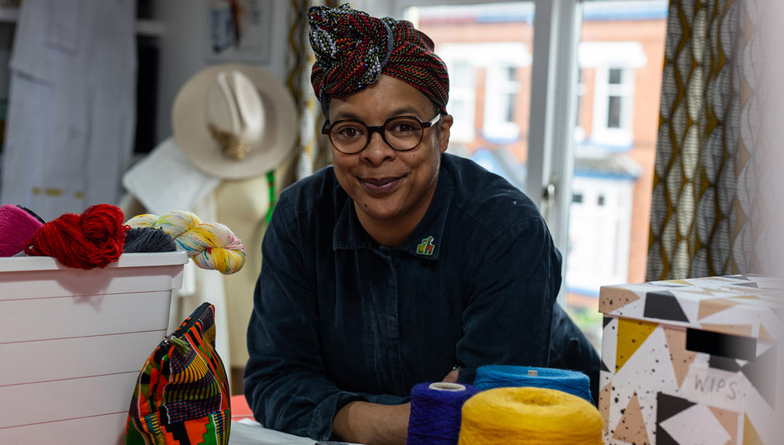 a Black woman rests her elbows on a table in a studio and looks at the camera. There are spools of thread, a box of yarn and other textiles and tools around her.