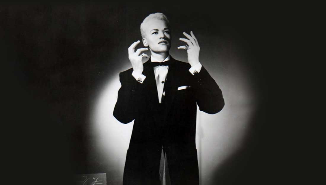 Black and white photograph, interior. Image of a male impersonator. They are wearing a suit blazer and bow tie, have short hair and are holding both hands up in front of them. They are not looking directly at the camera, but somewhere in the middle distance.