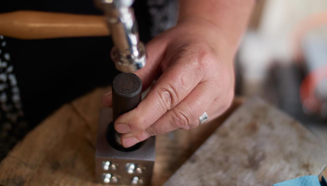 A hammer is visible above a hand holding a component into a metal cube with circular forms on its sides.