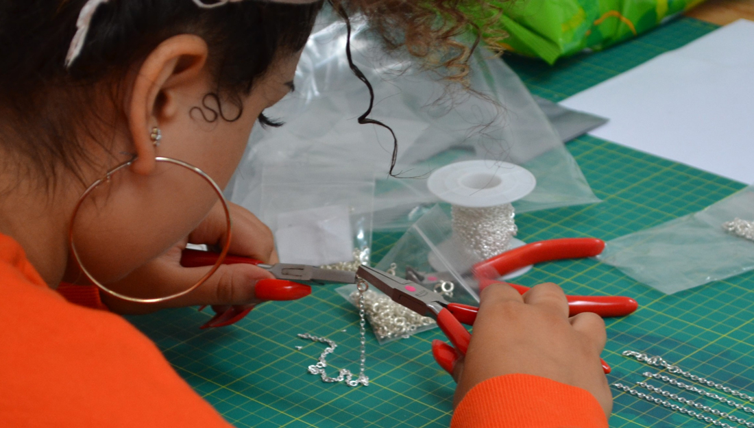 A young woman sits at a workbench working on a silver chain with pliers.