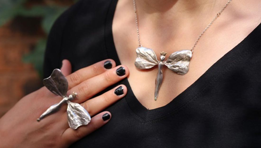 close up image of a silver necklace in the shape of a butterfly worn above a black v neck top. A hand wearing a large silver butterfly ring rests on the chest near the necklace.