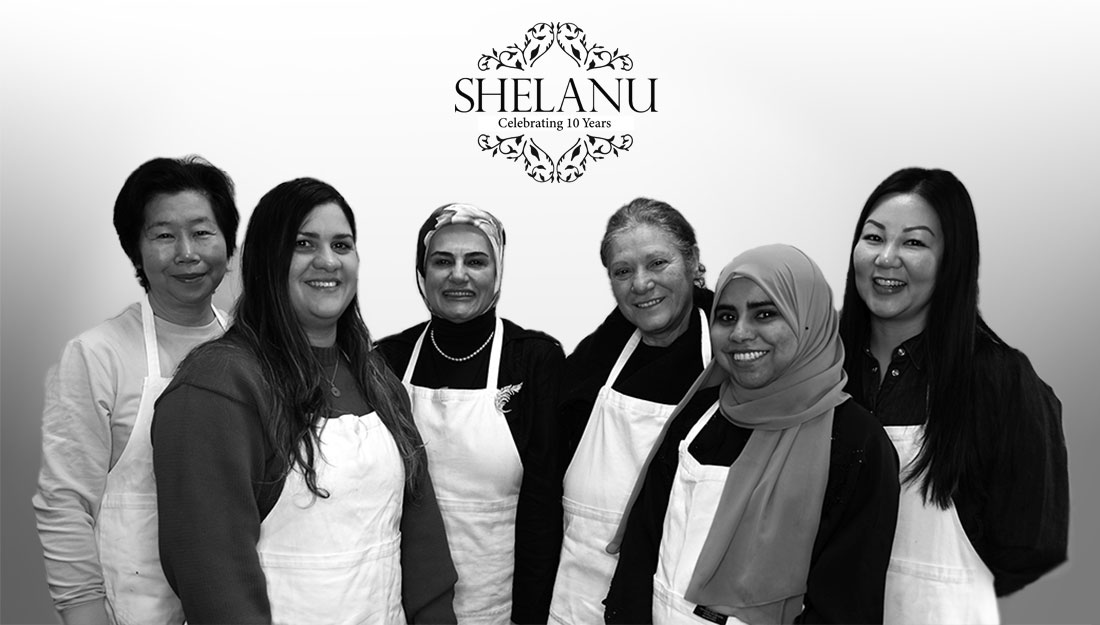 A group of 6 women from varied ethnic backgrounds and ages pose in white workshop aprons, they are all smiling.