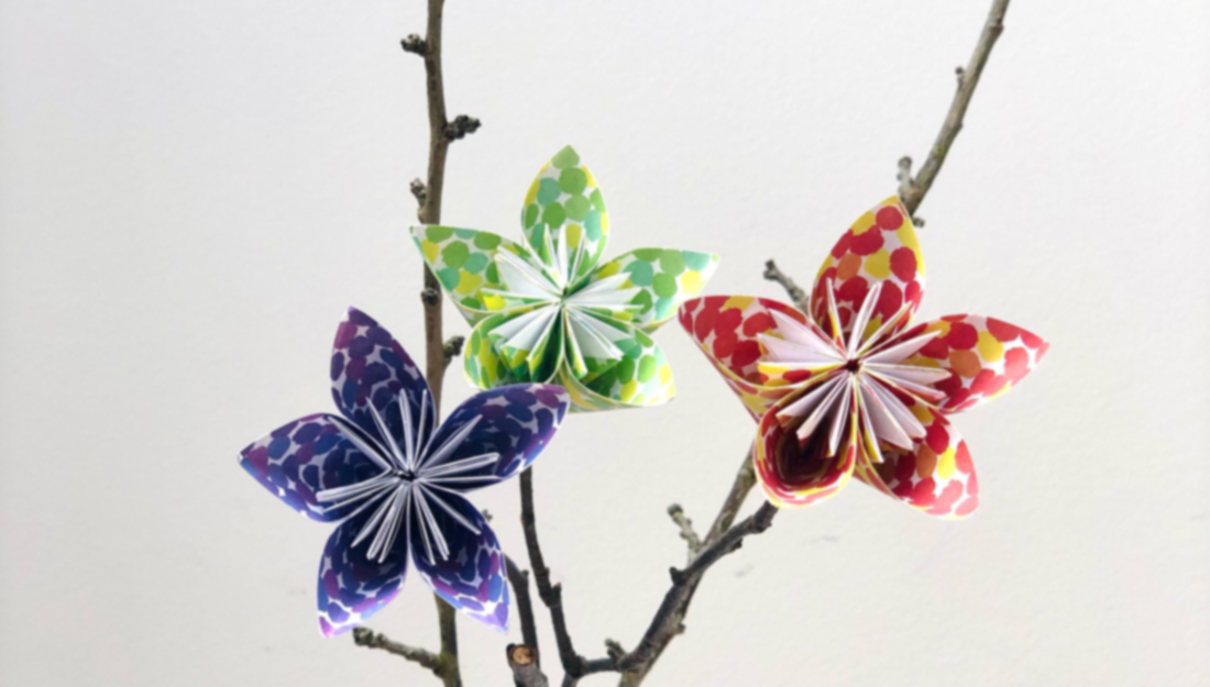 Three small pretty paper flowers, red green and purple, are displayed on twigs.