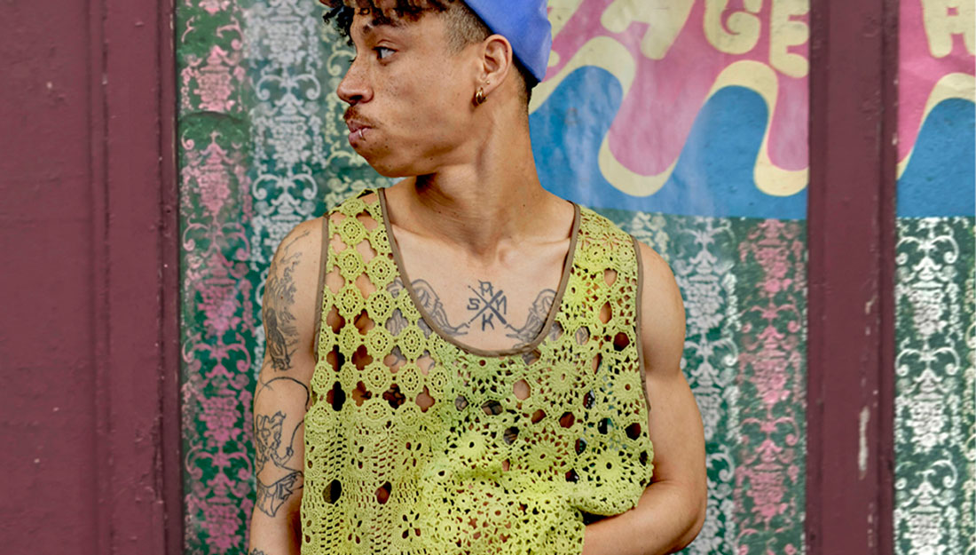 A fashion photo. A male model wears a baggy vest made from yellow lace and a blue beany hat. He has tattoos.
