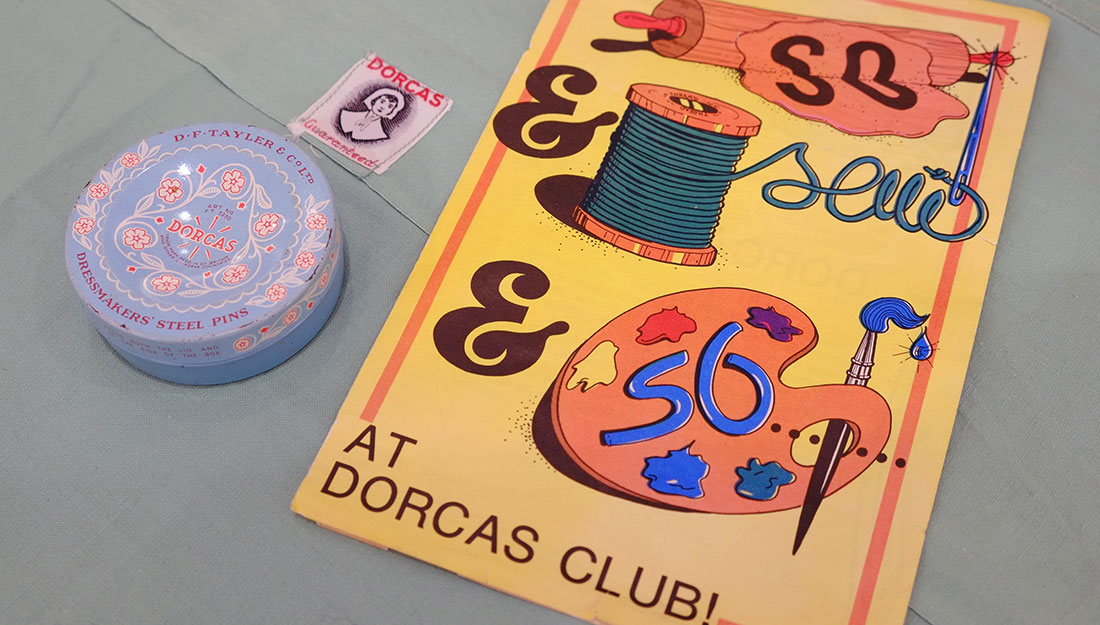 A bright vintage magazine with an illustrated cover showing a cotton reel, paint palette and rolling pin. Along side are a Dorcas label on a sewn item and a pin tin branded Dorcas.