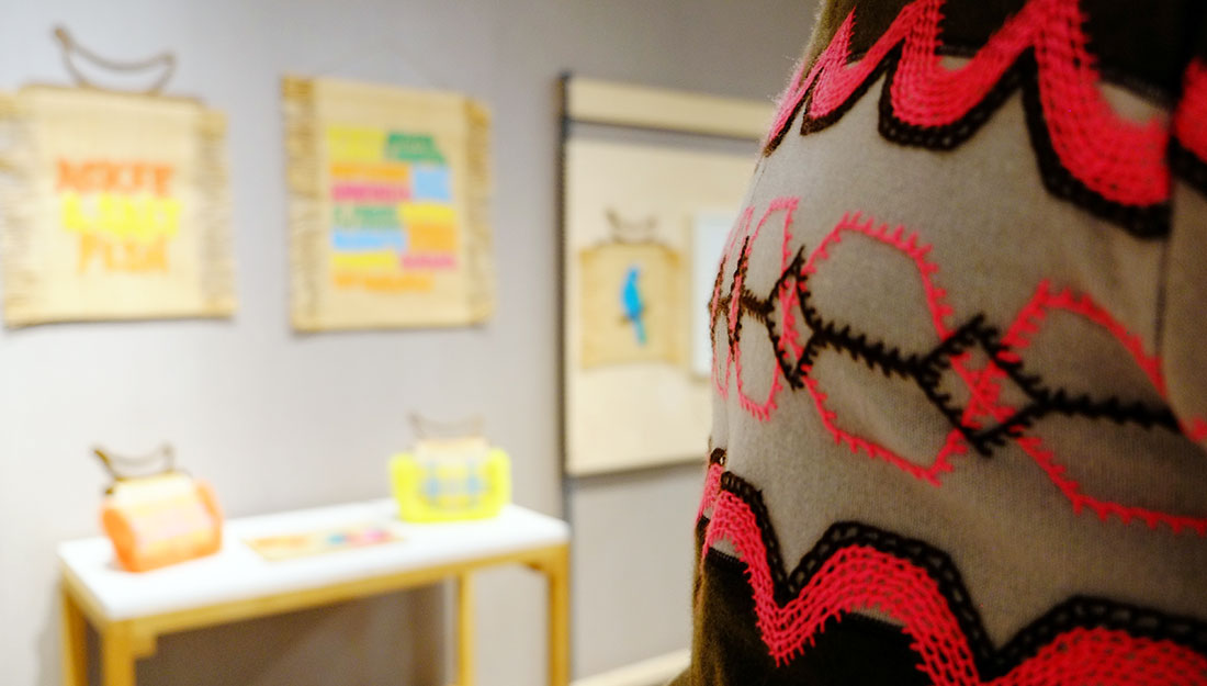 In the foreground a brown and grey knitted garment is embroidered with bright pink wool. The blurred backround shows some bright wall hangings.