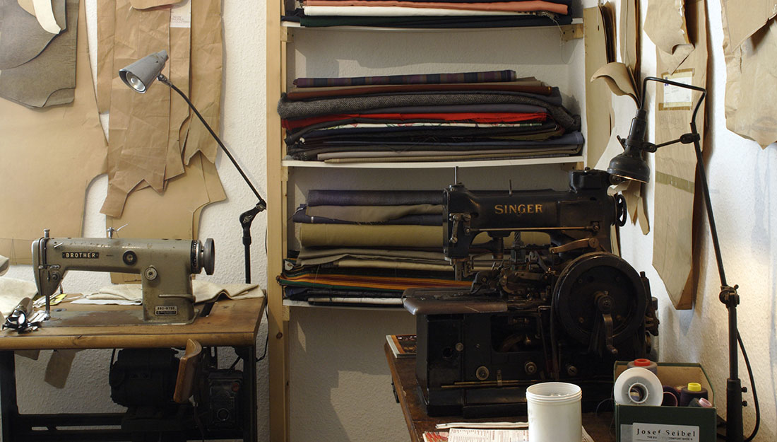 A cramped but drab looking sewing room. There are shelves of rolled up cloth, paper pattern pieces hanging on the walls and two sewing machines.