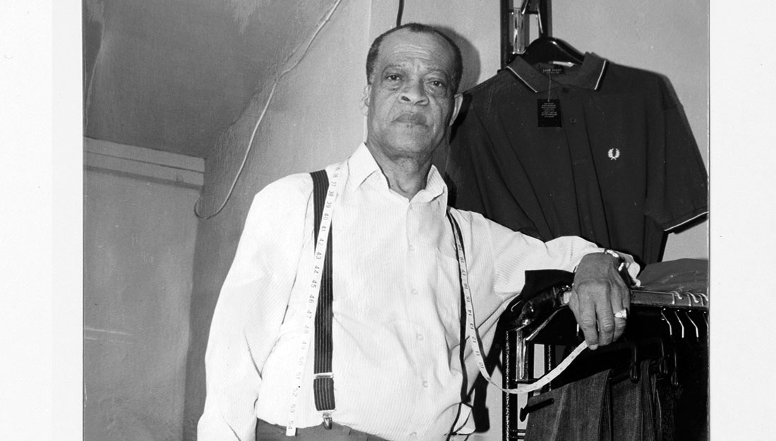 A portrait of an older black man. He has a tape measuer around his neck, a white shirt and braces on his trousers. He leans on a rail of clothes.