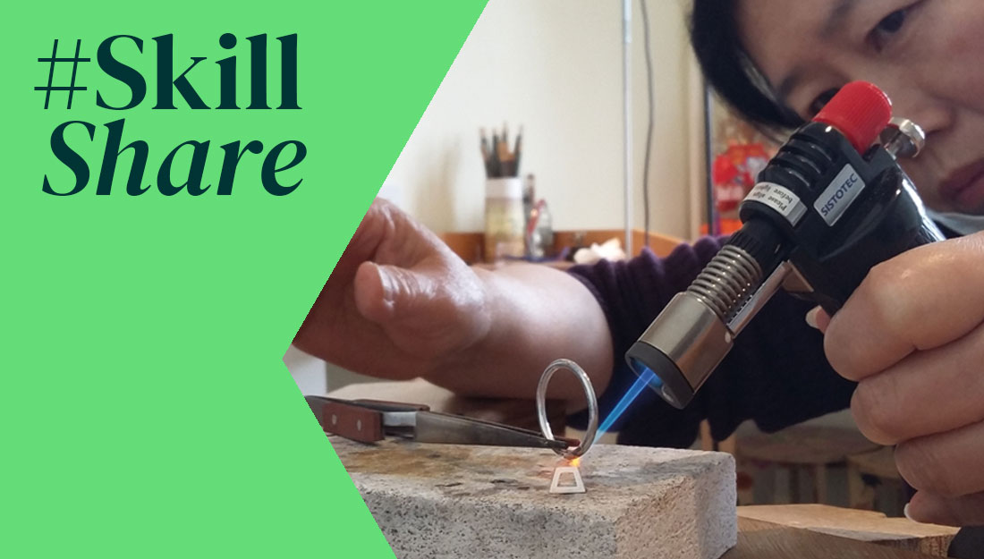 a close up of a ring being soldered on a heat brick using a blow torch. A woman's face is partially visible in the background. A green graphic with the words #SkillShare is overlaid over the top on the left.