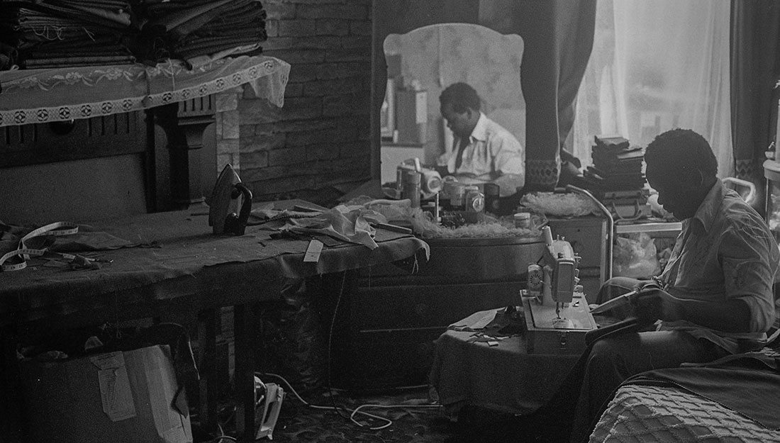 AN old black and white photo of a man sitting at his sewing machine in a cramped room. There is a small sewing table, a dressing table with a mirror, an iron on the floor and stacked rolls of fabric on the mantle piece.