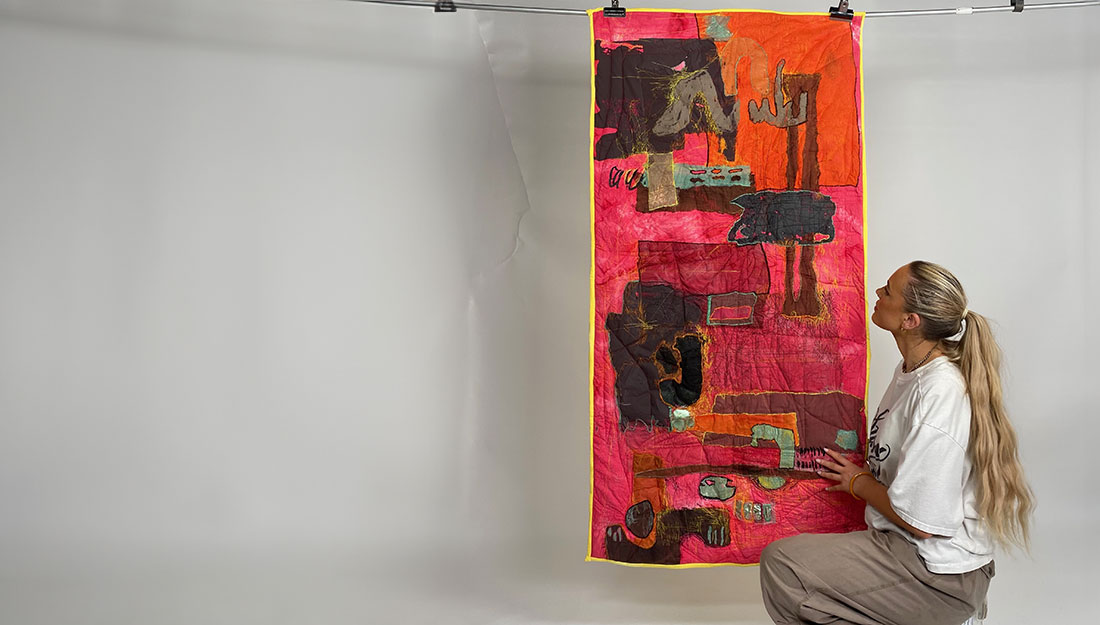 A woman crouching next to a rectangular, colourful textile piece hanging from a metal rod. The wall hanging is primarily pink and orange, with brown, black and green abstract shapes stitched or embroidered over it; the shapes and the piece are outlined with yellow thread.