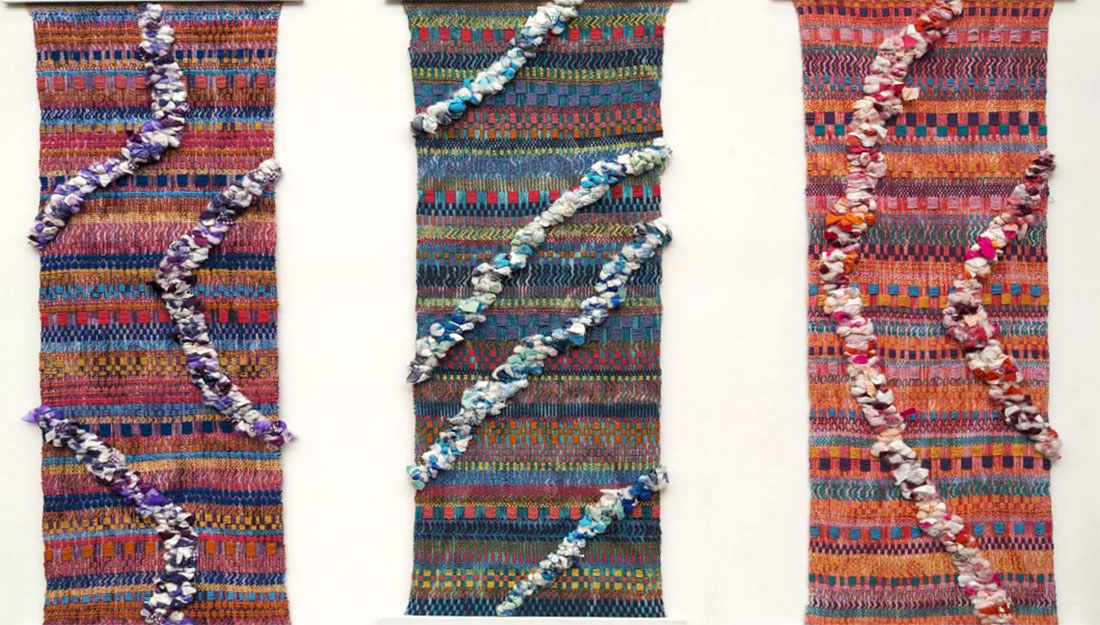 Three pieces of intricately coloured and designed cloth. The left-hand piece is largely pink, purple and orange; the middle piece is mostly green, turquoise, yellow, and red; the right-hand piece is pink, orange, and turquoise. These flat wall hangings have been decorated with sculptural curves that cut across them, made from scraps of similarly coloured fabrics.