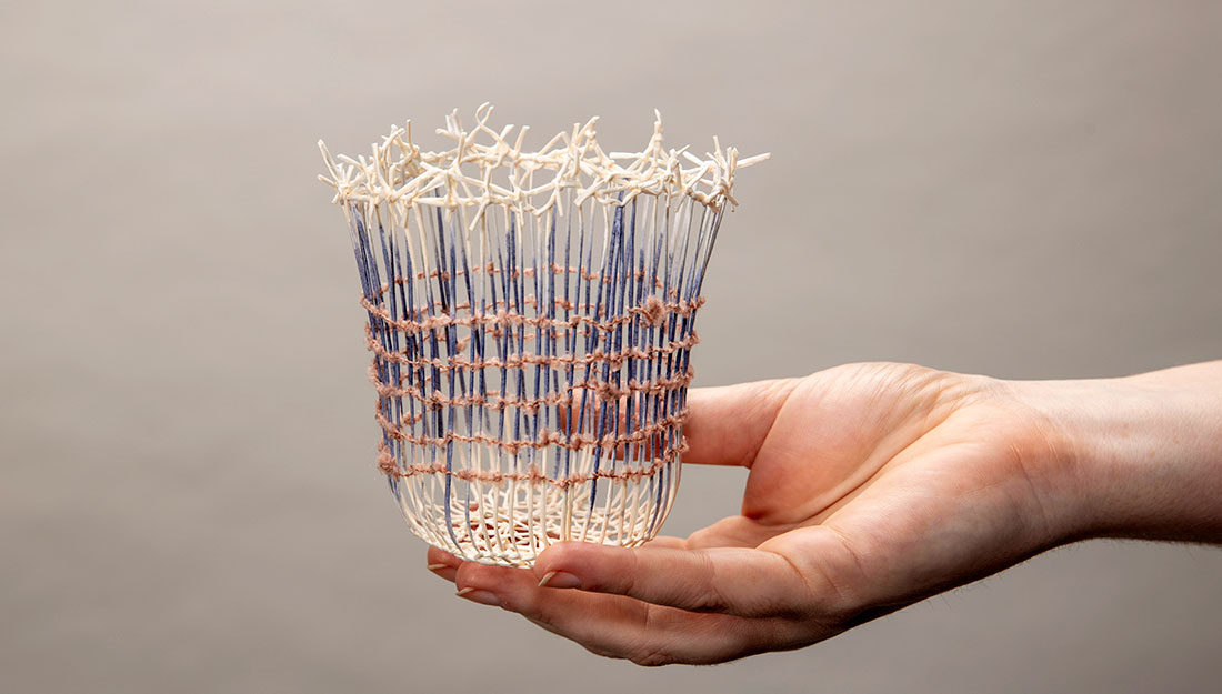 An open hand holding a very delicate cup-shaped object made from loosely woven, thin natural fibres. The vertical strands are blue and white, and the horizontal ones are pink. You can see through the vessel, almost like bars on a window. The joining at the top of the vessel appears like a crown of thorns.