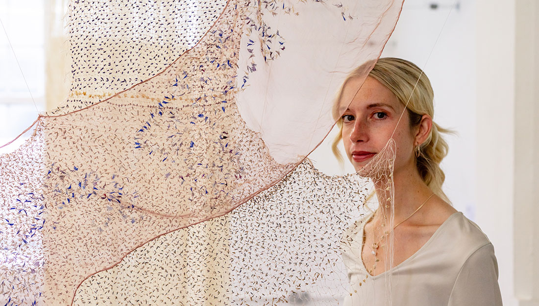 A blonde woman is a white t-shirt, wearing simple gold jewellery, stands just behind two overlapping stretches of translucent fabric, each embroidered with tiny flowers. The one running diagonally is a very thin, delicate pink chiffon decorated with tiny blue flowers; the other runs more vertically – a delicate web of incredibly thin natural fibres embroidered with tiny flowers and petals.