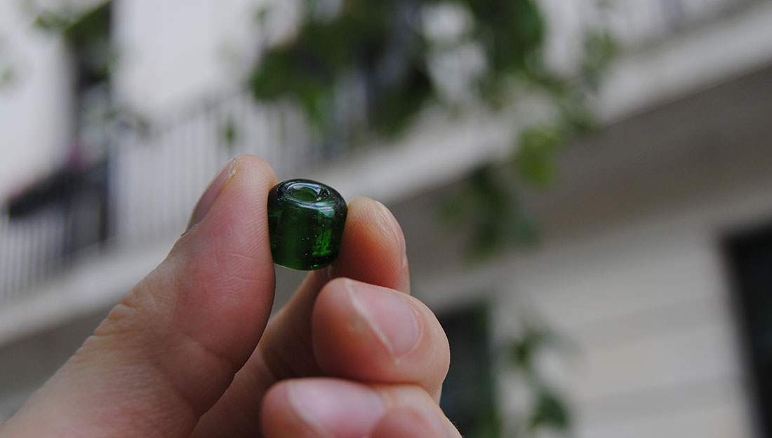 A hand holding a green glass bead between the thumb and forefinger in front of a blurred house and tree.