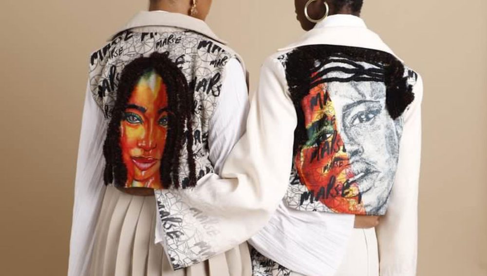 Two women, dressed all in white, stand with their backs to us, and their arms around each other’s backs. The backs of their jackets have been decorated with colourful portraits of Black women’s faces, with their hair embroidered on.