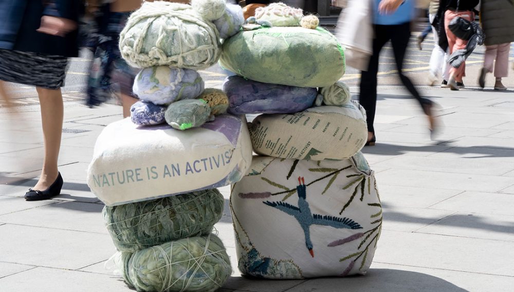 An arch constructed out of different-sized fabric boulders, in the middle of a busy pavement. The largest boulders have been embroidered and others printed on – one has a crane swooping down, one says ‘Nature is an Activist’.