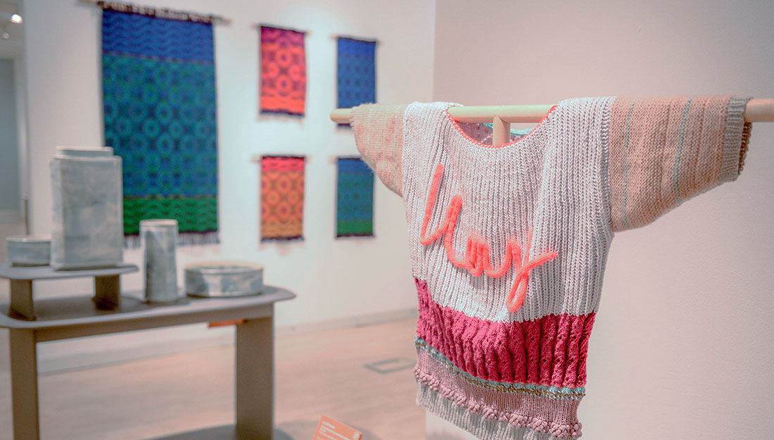 A view of an exhibition in a gallery. In the foreground is an embellished knitted jumper on a frame. Behind are ceramic vessels and on the wall are bright woven wall hangings.