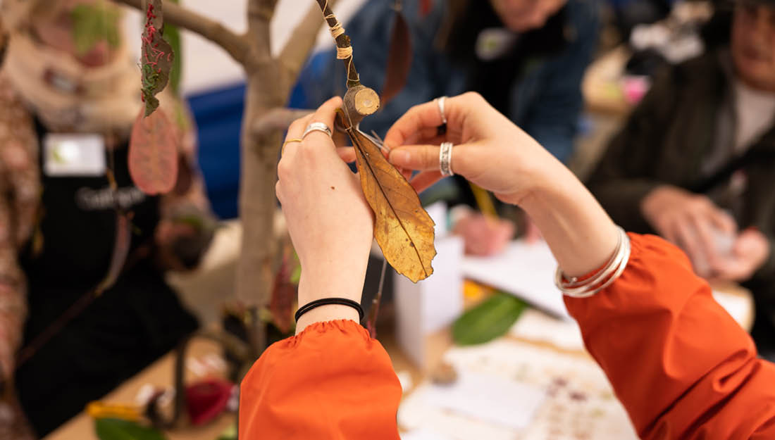 A close up of hands tying a yellowed, embroidered leaf onto a bare branch, next to other hanging embroidered leaves.