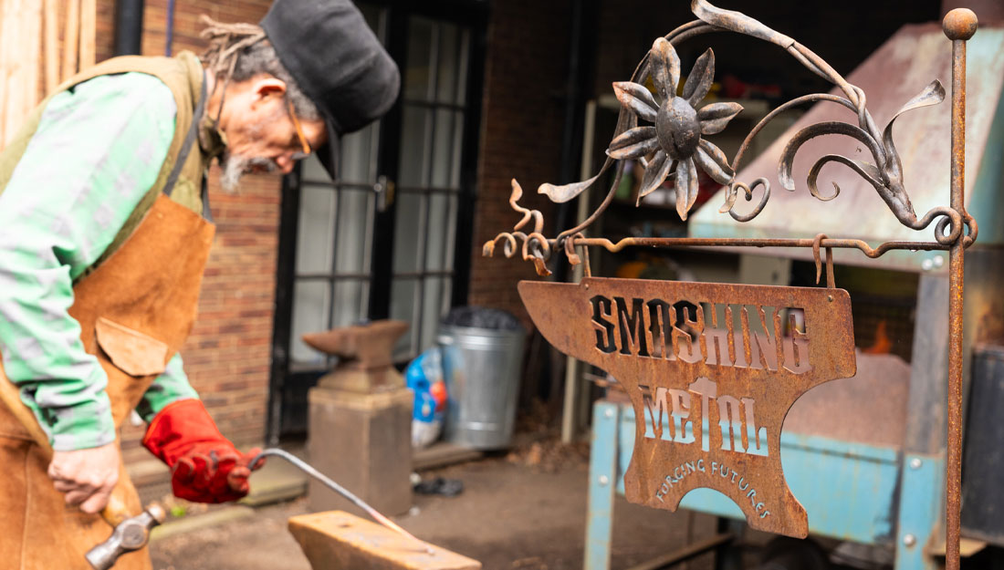 Alex, the leader of Smashing Metal, wearing an apron and heatproof gloves, holding a hammer in his left hand and a hot metal rod in his left, shaping it against an anvil. In front of him is a metal sign shaped like an anvil, with letters cut out which reads 'Smashing Metal Forging Futures'. The rod the sign hangs from has been decorated with a metal flower and stem which arches over the stop of the sign.