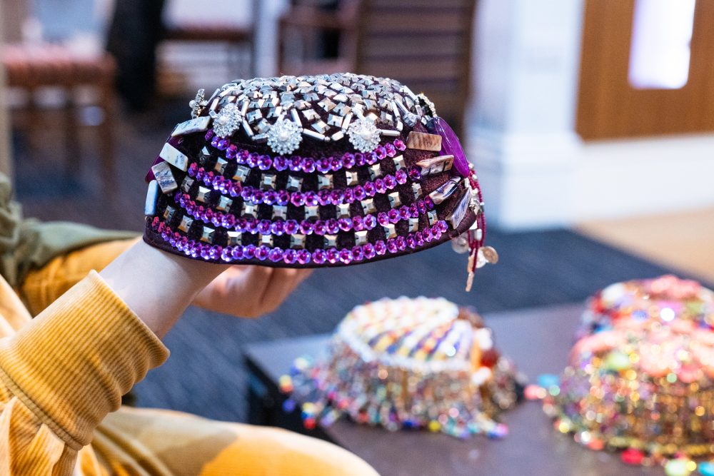 A brown velvet Kurdish woman's cap made by Zohra, embellished with silver and purple sequins and beads, being held up above the table where the rest of the caps are laid out.