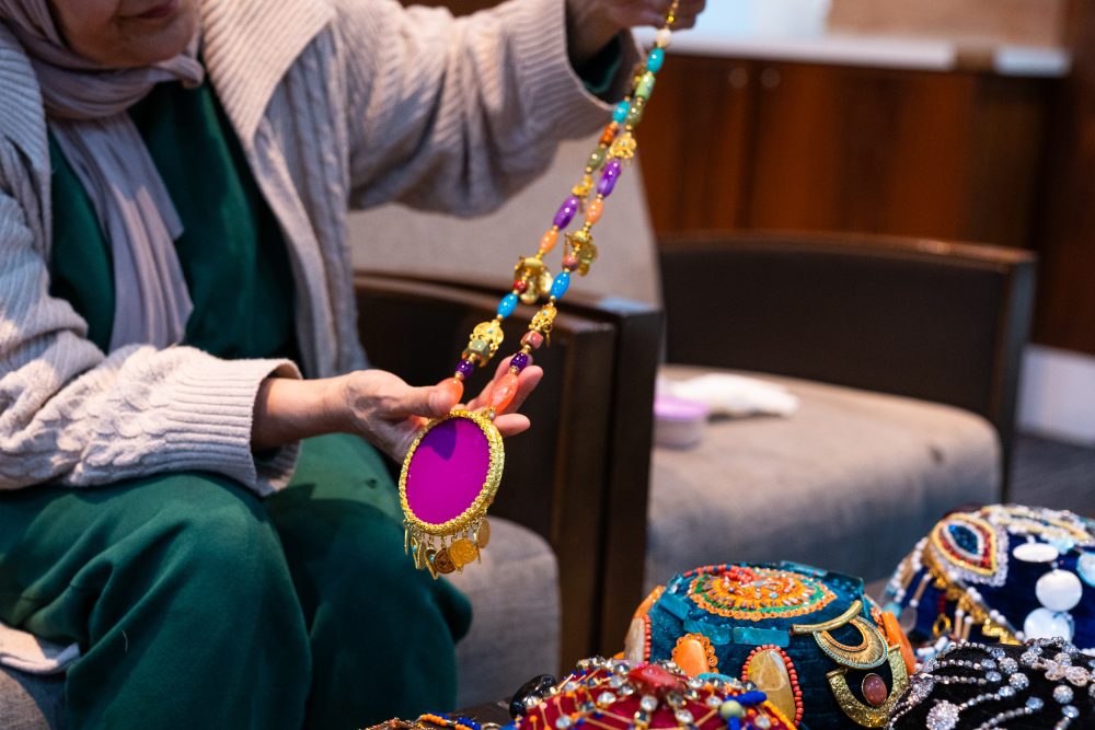 A woman dressed in green and pink, holding up a beaded necklace with a large pink pendant.