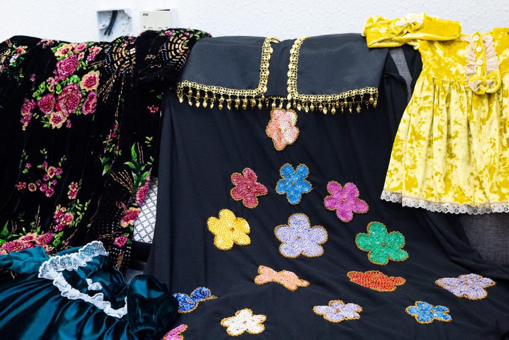 Garments handmade by Zohra laid out on a chair. There are children's dresses, jewel-tone in colour and finished with white lace trims. There are also two black garments embellished with beadwork.