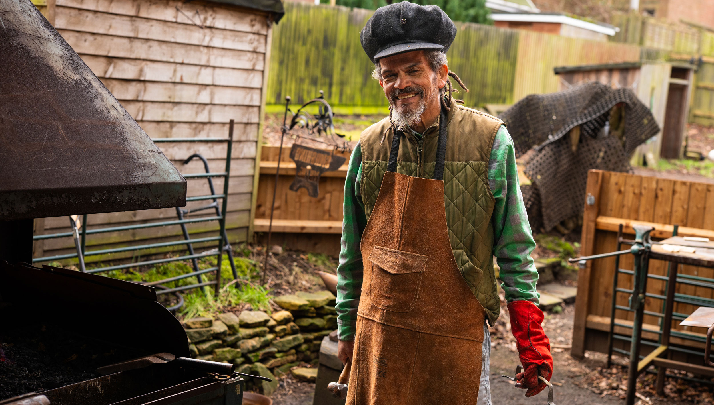 Alex, the leader of Smashing Metal, standing in the outdoor metalworking workshop next to the forge, with the anvil, the clamp table and the sign behind him. He is smiling and looking at the camera.
