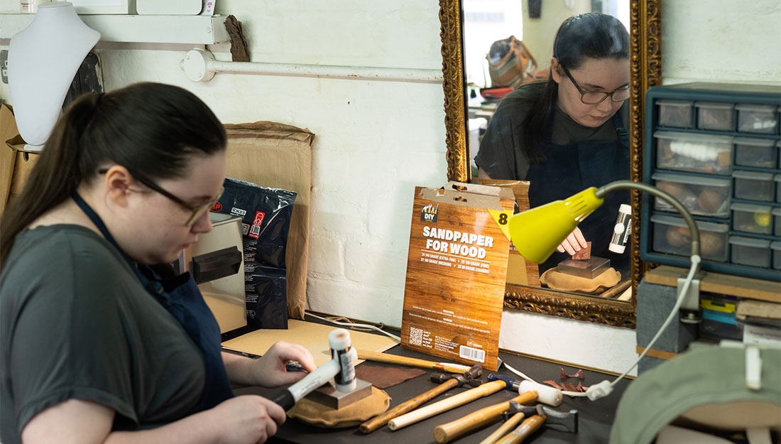 CraftingPathways_Megan&Nobuko_PhotoHayleySalter_3 Megan at a bench hammering a piece of metal, surrounded by jewellery working tools.